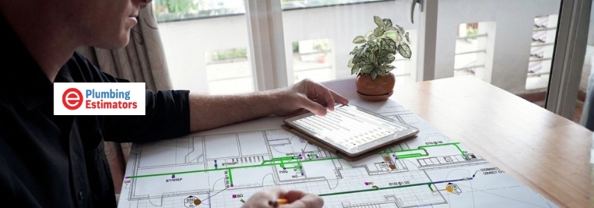 A plumbing estimator using a tablet to review project specifications over a detailed hydraulic layout blueprint on a desk.