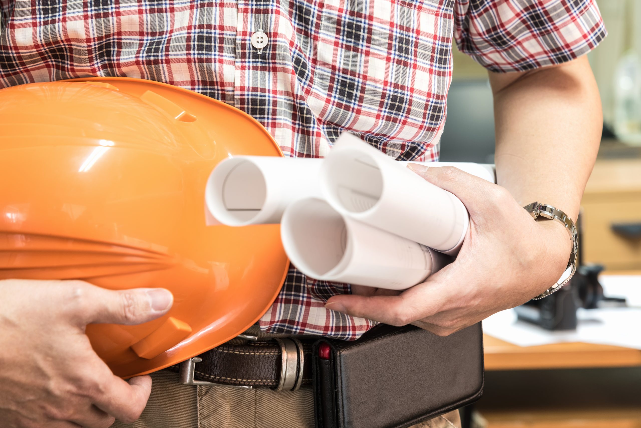 Close-up of a plumbing estimator wearing a plaid shirt and tool belt, holding an orange hard hat and rolled-up project blueprints to estimate site conditions for trench drain and concrete pit pricing.